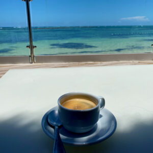 Coffee on a white table with ocean in the background. Nancy's summer blogging challenge