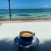 Coffee on a white table with ocean in the background. Nancy's summer blogging challenge