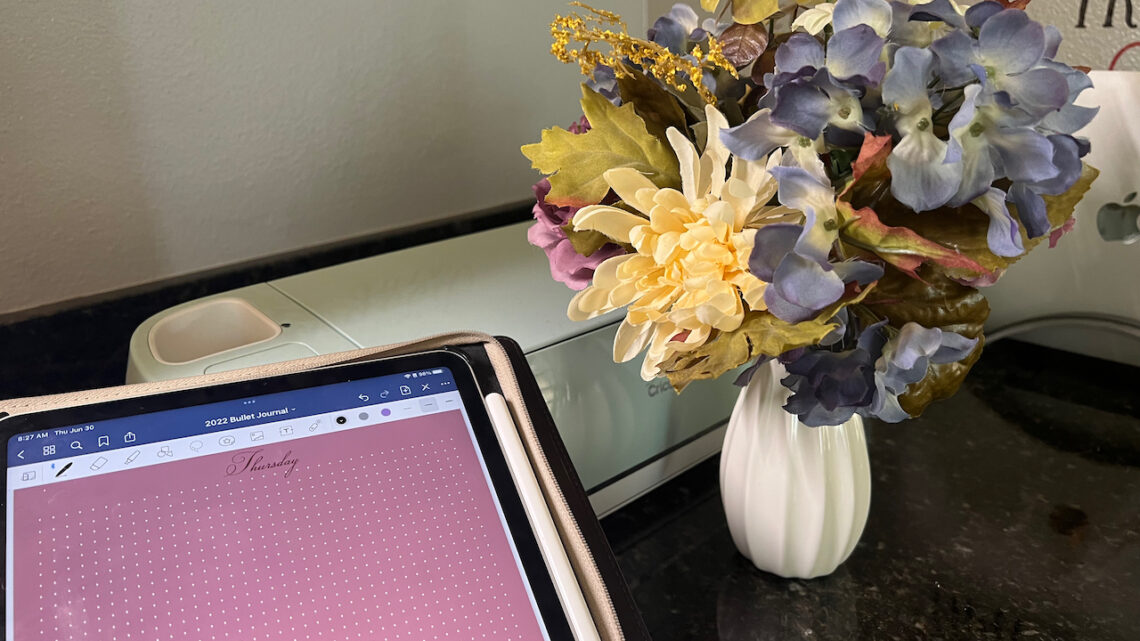iPad with pink screen on a desk with a craft machine and flowers. ho eI use GoodNotes stickers
