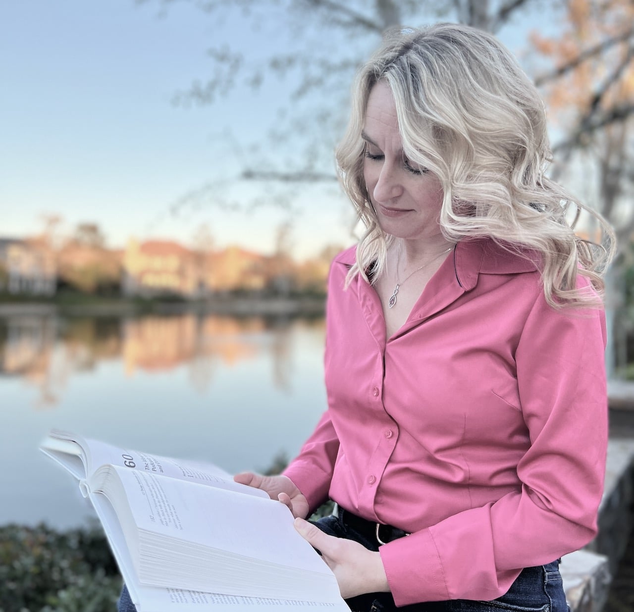Jennifer Douglas reading a book in a pink shirt sitting by a lake
