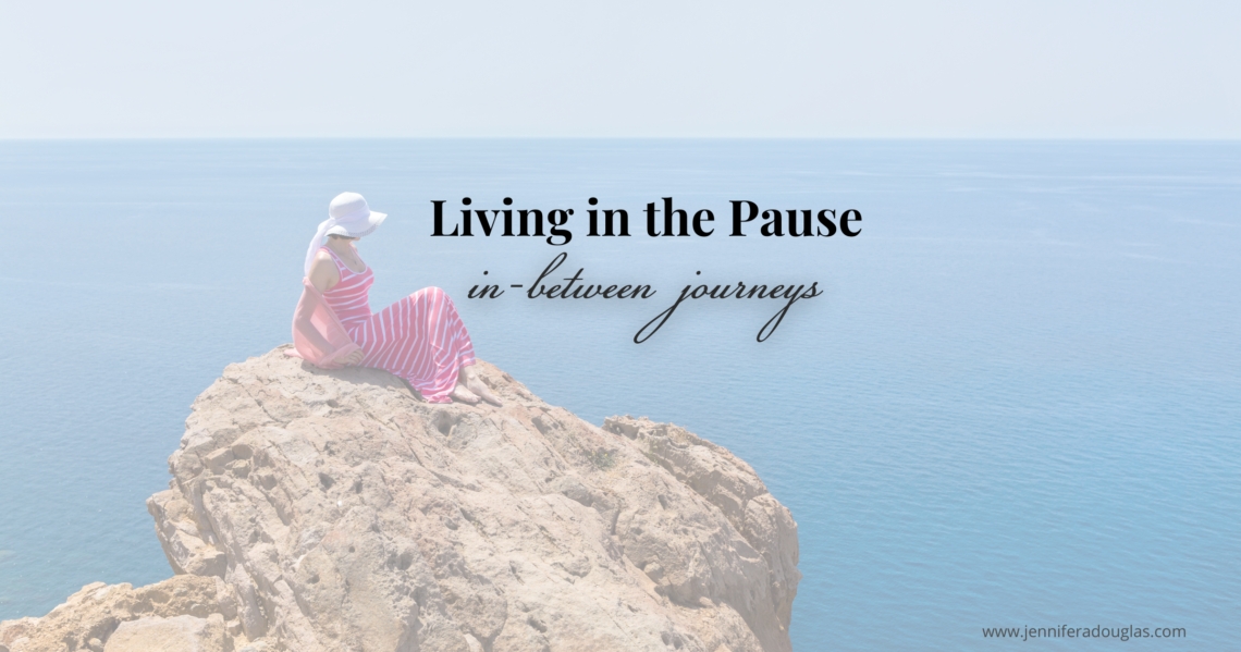 Photo of a woman in a pink dress sitting on a rock overlooking the ocean. Text reads Living in the Pause: In-between journeys